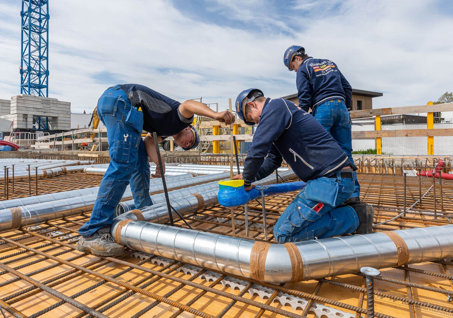 Industriefotograf Luzern: 3 Männer in blauer Arbeitskleidung auf montieren silberne Alurohre auf einer Aussenbaustelle