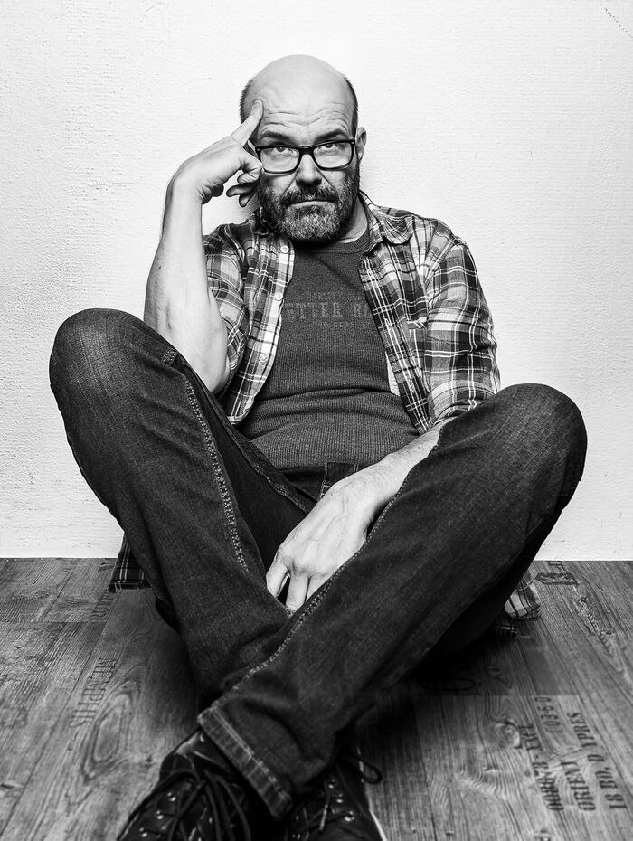 Patrick Wicki – Mann in denkender Pose an Wand sitzend auf Holzboden mit orangefarbener Typoschrift